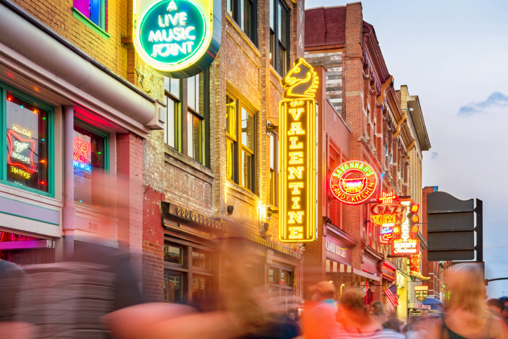 Stock photograph of people and colorful neon signs in a row at the landmark Broadway pub district in downtown Nashville, Tennessee, USA, illuminated at twilight blue hour